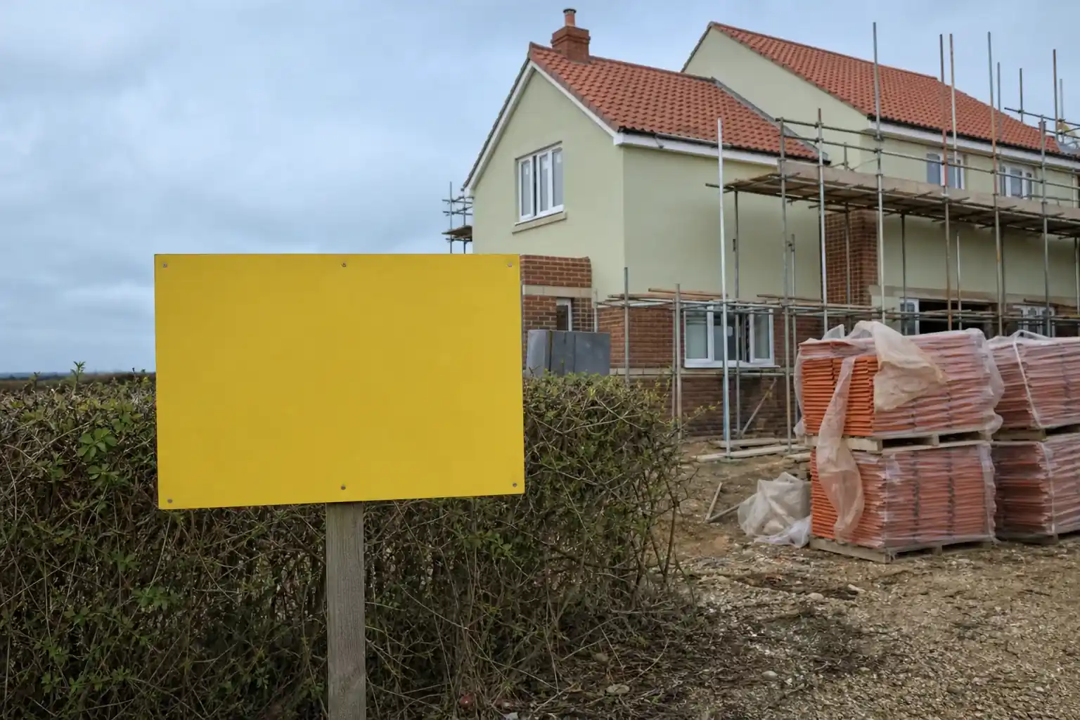 Panneau d’affichage de permis de construire vierge planté en limite de terrain, maison en construction en brique et enduit à l’arrière-plan