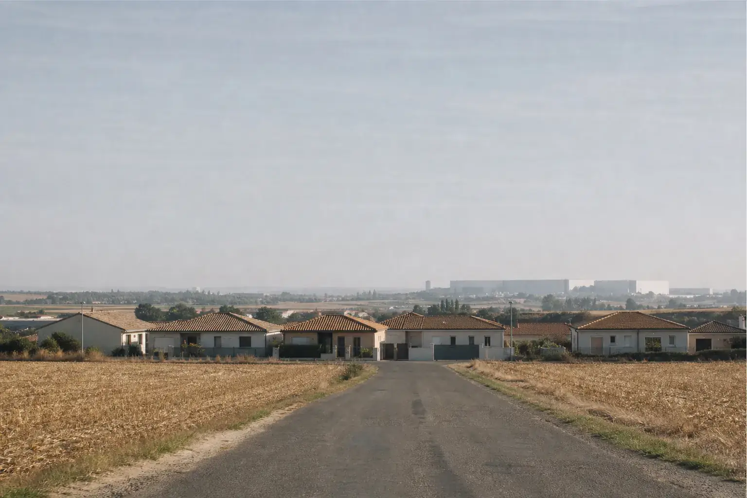 Terres agricoles planes en lisière de village au nord-ouest de Toulouse, route droite menant à des maisons neuves, immense hangar aéronautique à l’horizon