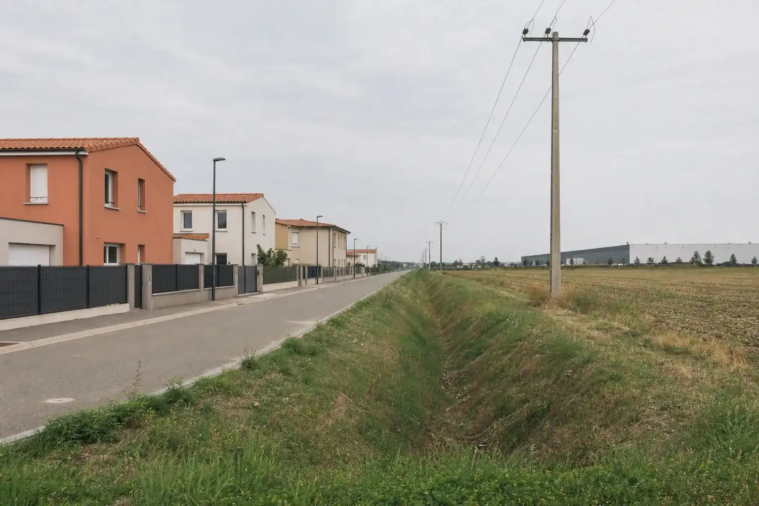 Rue résidentielle en limite nord de l’agglomération toulousaine, maisons neuves d’un côté, champ ouvert et entrepôt lointain de l’autre