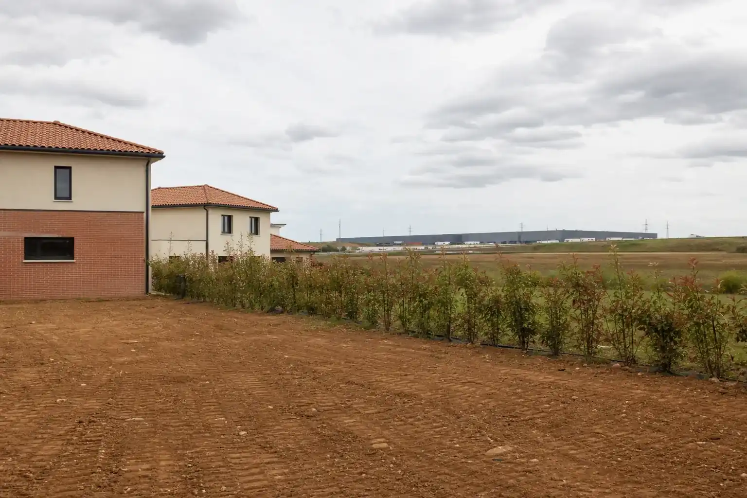 Terrain en limite nord de l’agglomération, maisons neuves d’un côté, plateforme logistique et talus autoroutier au loin, haie de protection en limite