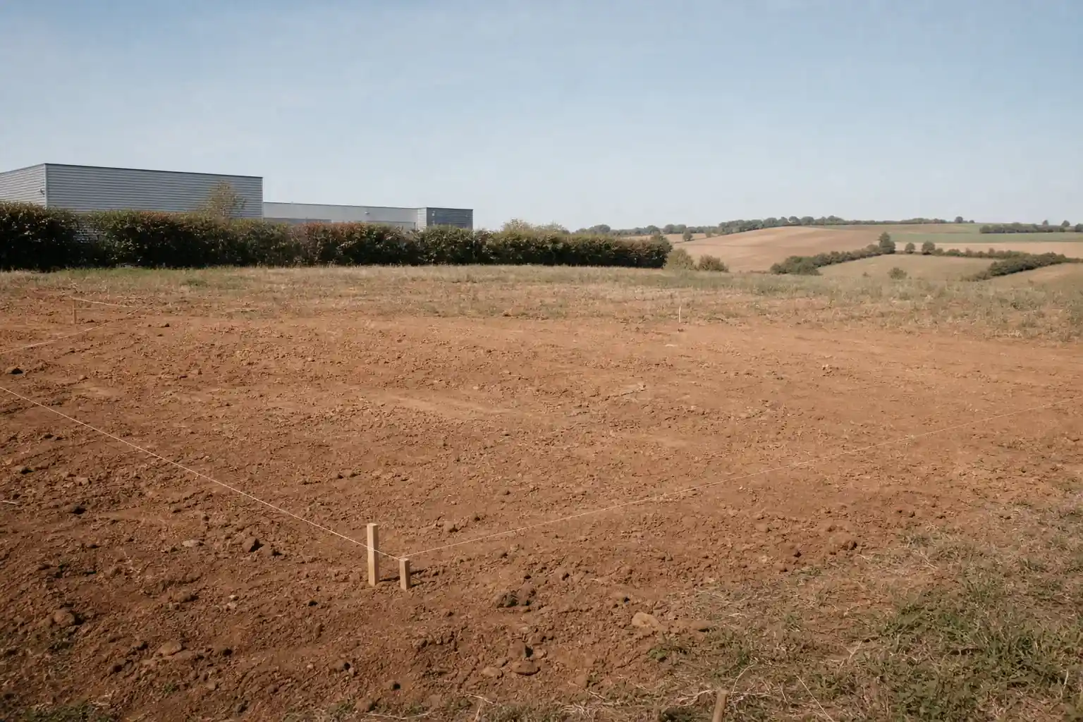 Terrain à bâtir fraîchement piqueté en lisière de bourg, entrepôts d’une zone d’activité derrière une haie, champs vallonnés de l’autre côté