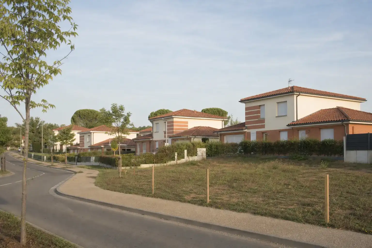 Lotissement récent sur un coteau au sud-est de Toulouse, rue courbe, maisons en retrait derrière des haies basses, une parcelle encore libre