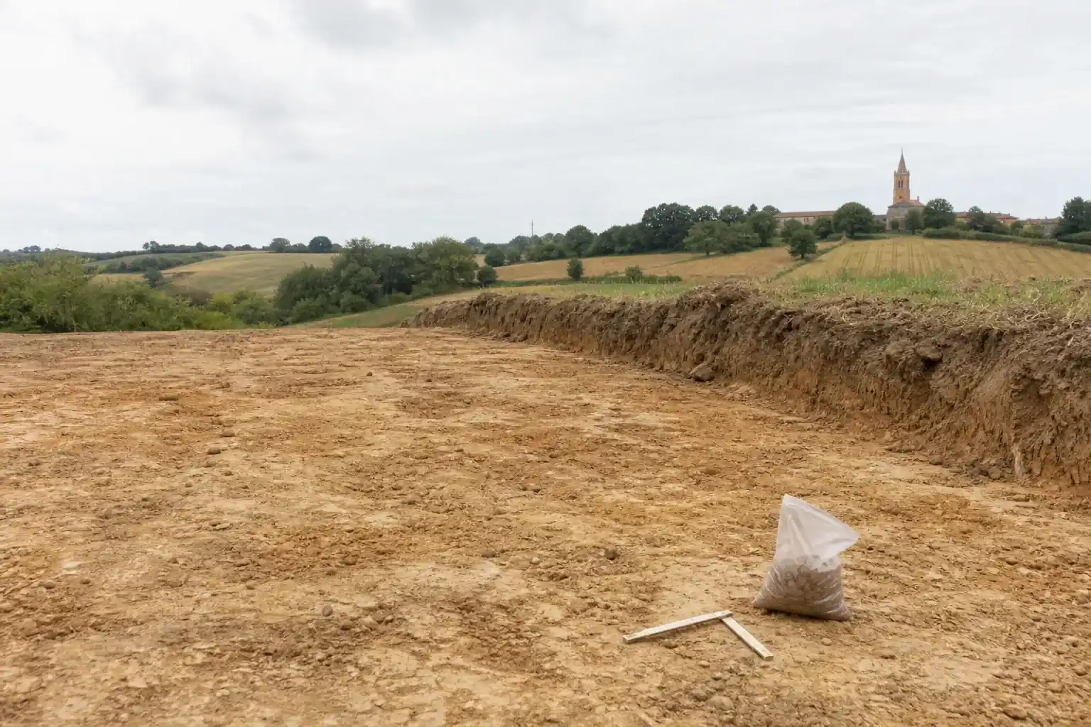 Terrain décapé sur un relief doux au sud-ouest de Toulouse, sous-sol limono-argileux brun clair mis à nu, sachet d’échantillon posé au sol